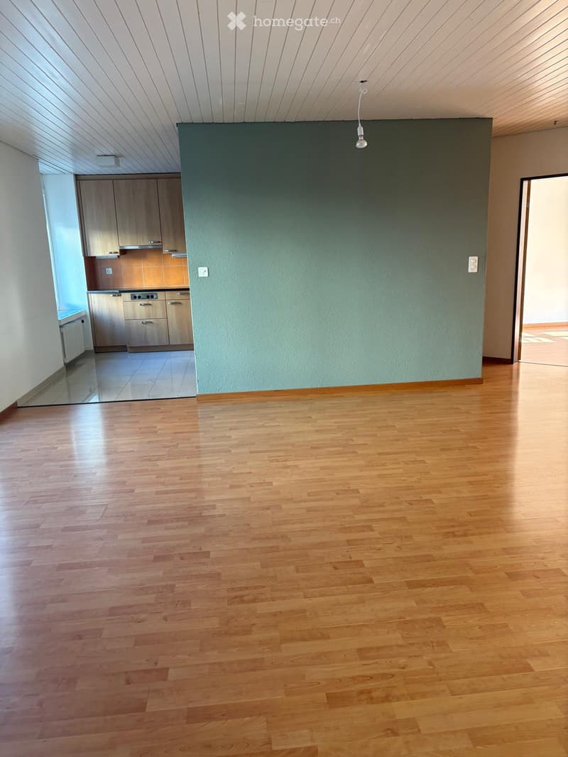 Empty room with wooden flooring, light green wall, and a small kitchen area visible in the background.