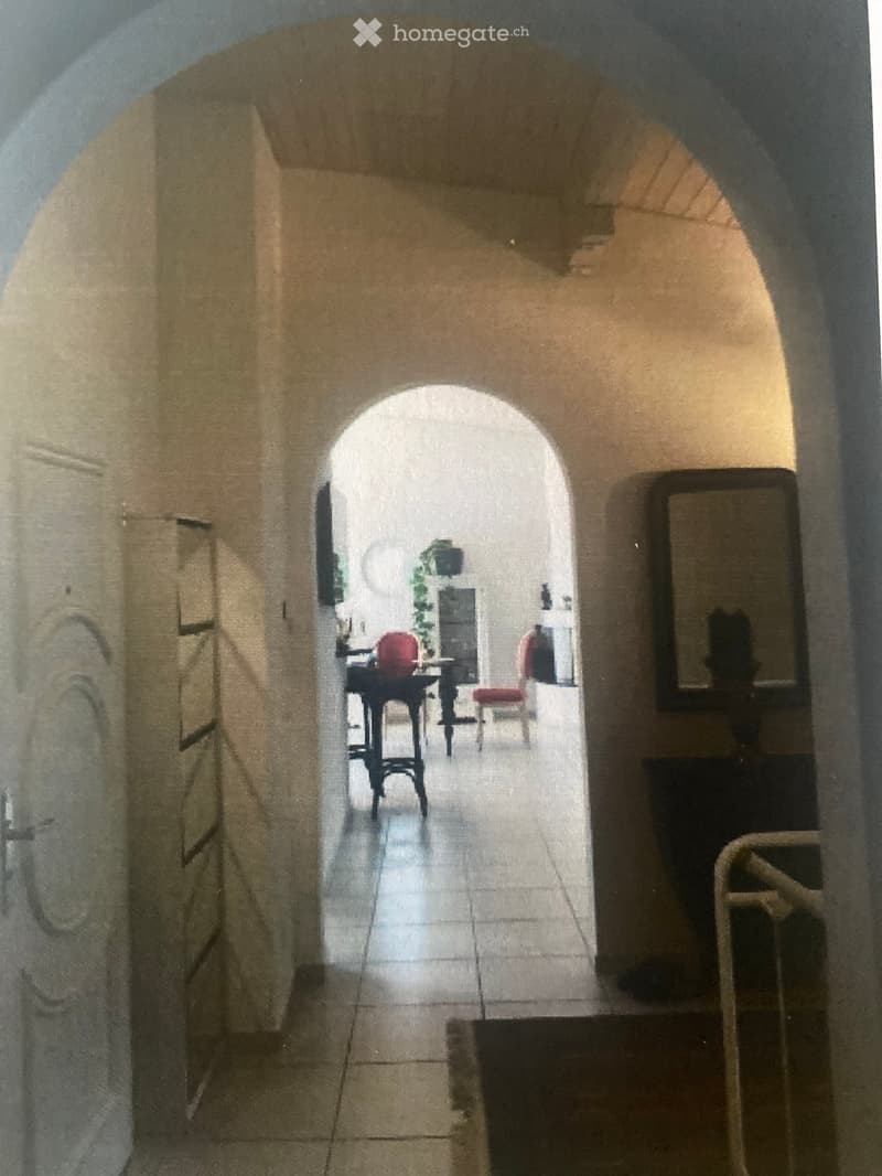 A narrow, arched corridor with tiled flooring and beige walls, leading to a room with a small table, chairs, and a mirror on the wall.
