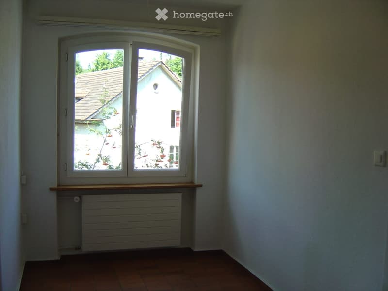 Empty room with wooden flooring, white walls, a large window with a view of a neighboring house, a radiator below the window, and a small wooden shelf above the radiator.