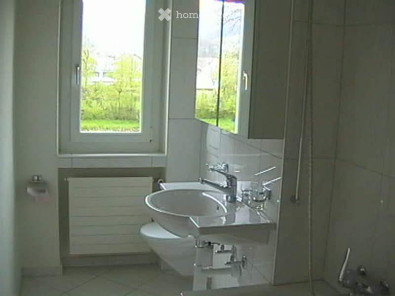 Modern bathroom with white tiles, a sink, toilet, mirror, and radiator. Natural light from a window illuminates the space.
