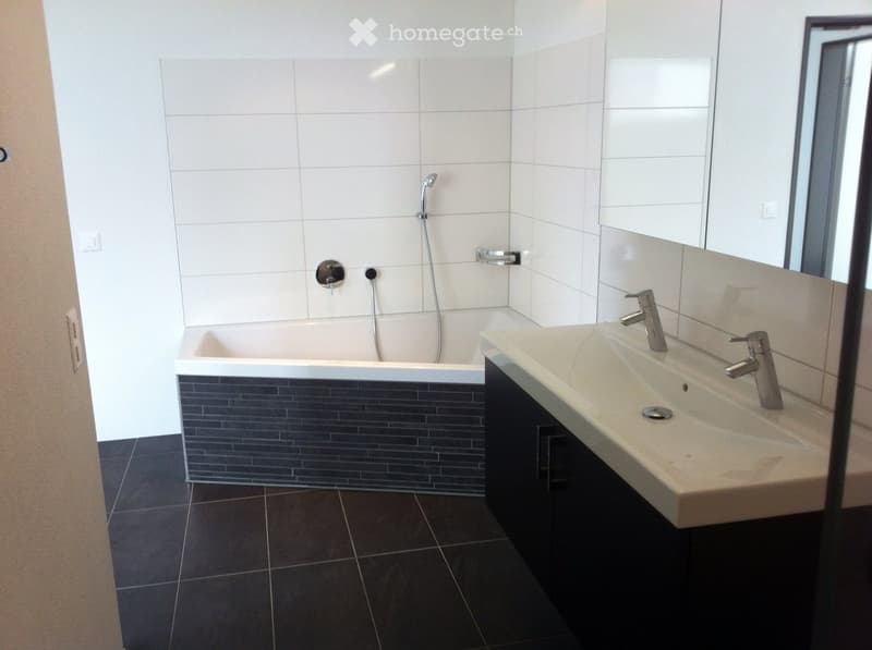 Modern bathroom with a bathtub, sink, and chrome fixtures. The walls are tiled in white and grey, and the floor is dark tiled.