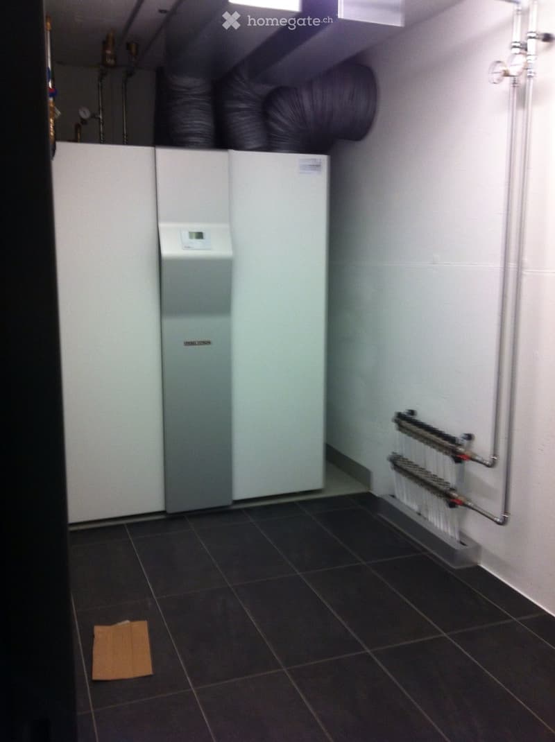 Utility room with tiled floor, white walls, heating unit, water tank, and pipes. No furniture or personal items present.