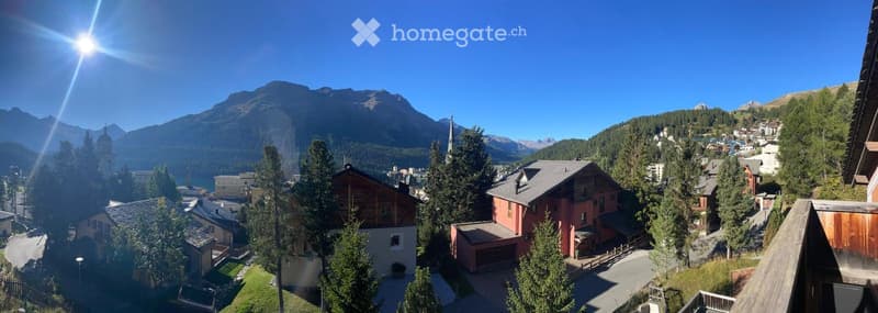 Panoramic view of a village nestled in a mountainous landscape, with houses, trees, and a clear blue sky.