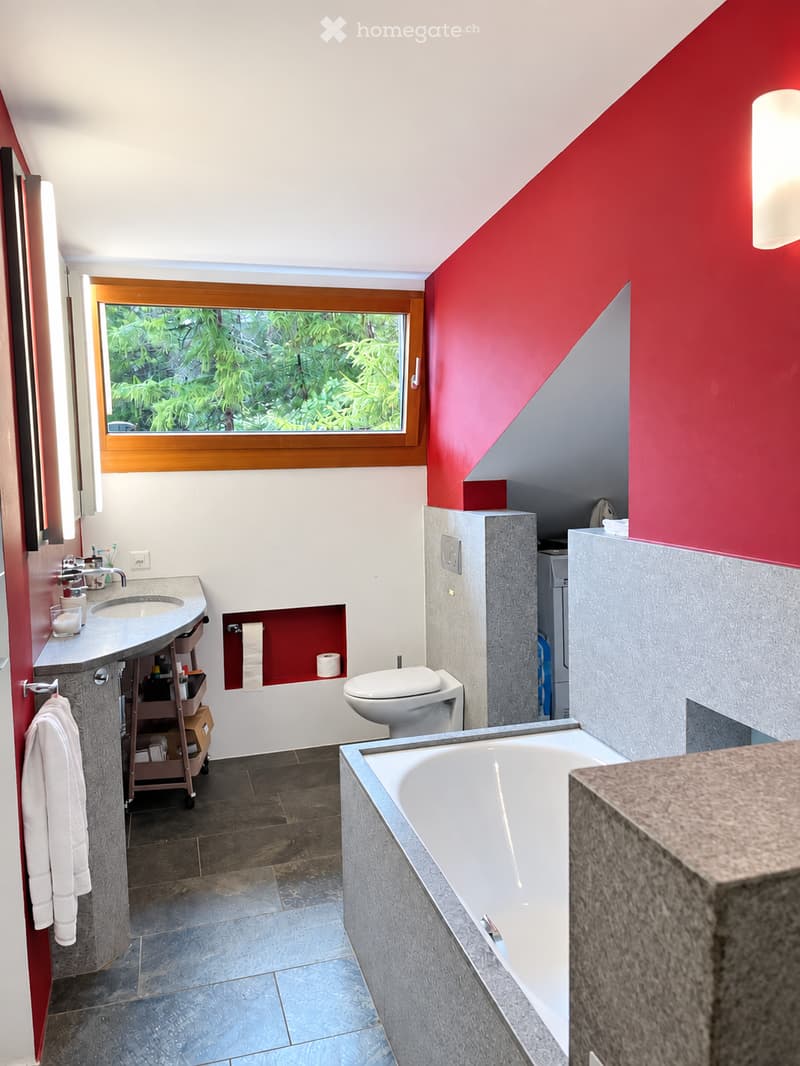 Modern bathroom with a red accent wall, white bathtub, toilet, sink, and a large window overlooking greenery.