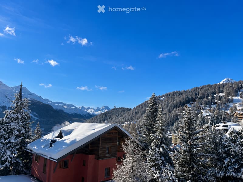 A red wooden house with a snow-covered roof, surrounded by snow-laden trees and mountains in the background.
