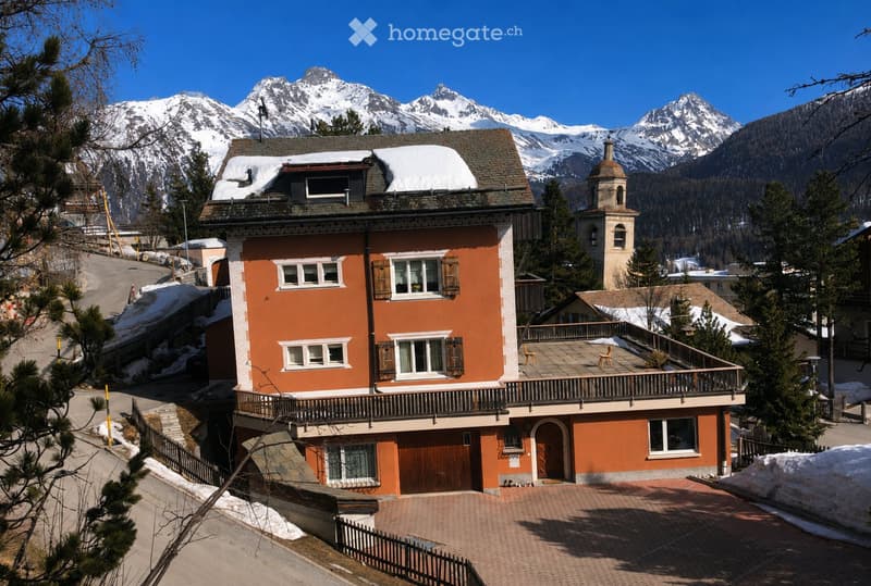 A two-story orange house with white trim, multiple windows, and a balcony overlooking a snow-capped mountain range.