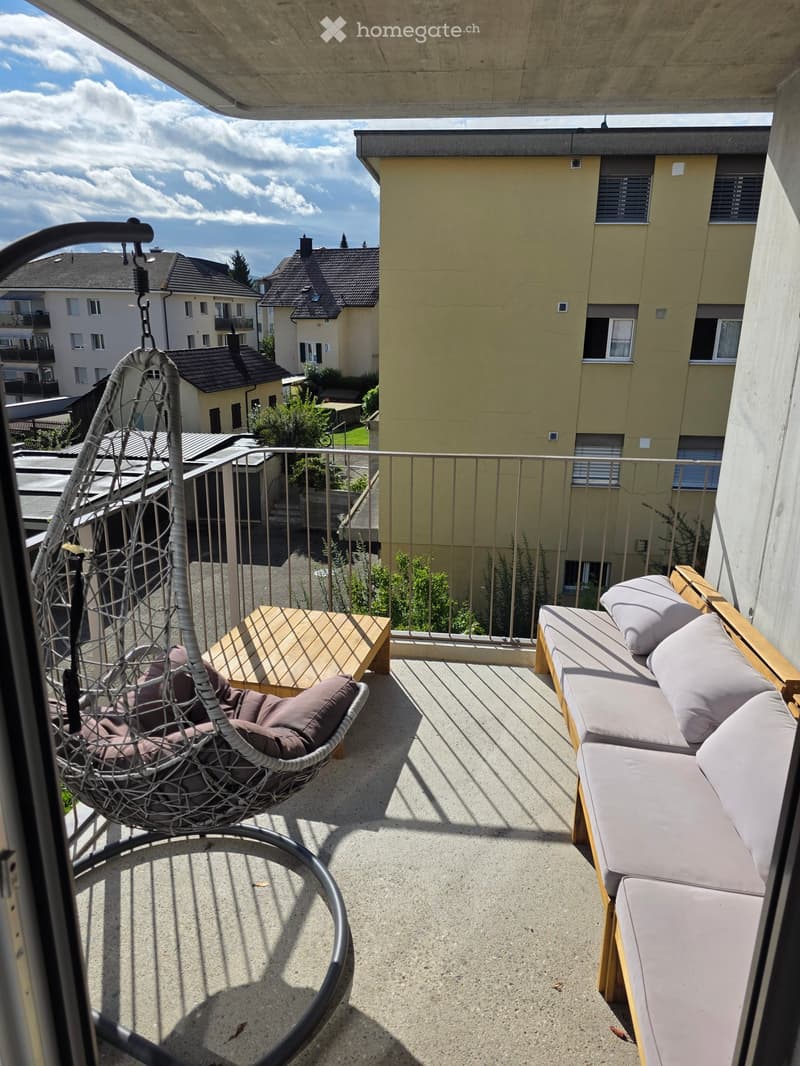 Balcony with a modern sofa, small wooden table, and a wicker hanging chair, overlooking a residential area.