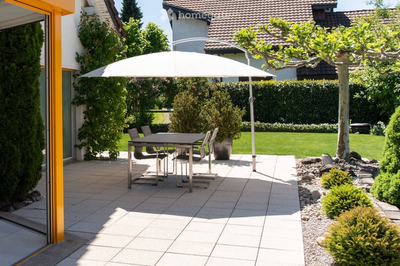 A well-maintained garden patio with a large umbrella, a dining table with four chairs, and surrounding greenery including shrubs, trees, and a neatly trimmed lawn.