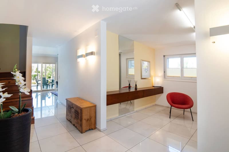 Modern hallway with white tiled flooring, a red chair, a wooden stool, a large mirror, a table with decorative items, and a potted plant near the staircase leading to an outdoor area.