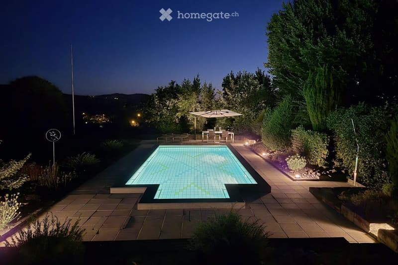 Well-lit outdoor area with a swimming pool, patio, outdoor dining set, umbrella, and surrounding greenery.
