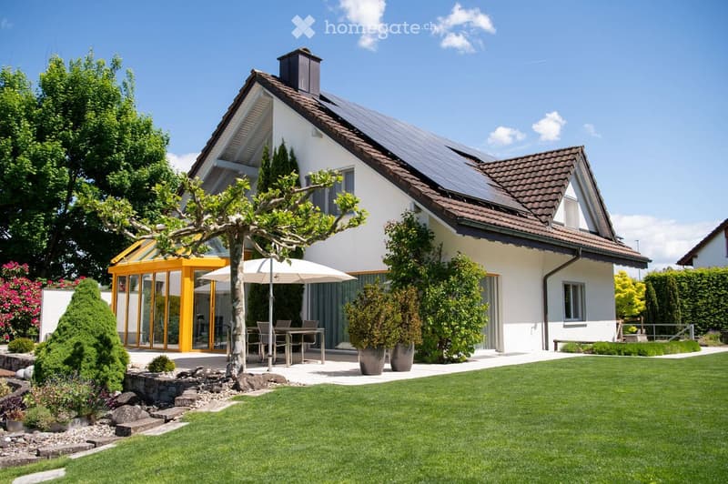 Two-story house with a modern design, featuring a tiled roof, large windows, and a spacious garden with a patio area, outdoor furniture, and a tree.