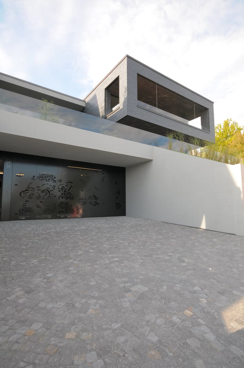 Modern two-story house with grey brick facade, large windows, and a raised ground floor. Features a glass balcony and decorative metal door.