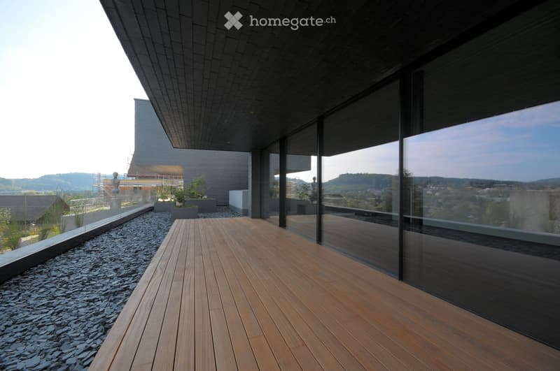 Modern wooden deck terrace with glass railings, overlooking a cityscape and distant hills.
