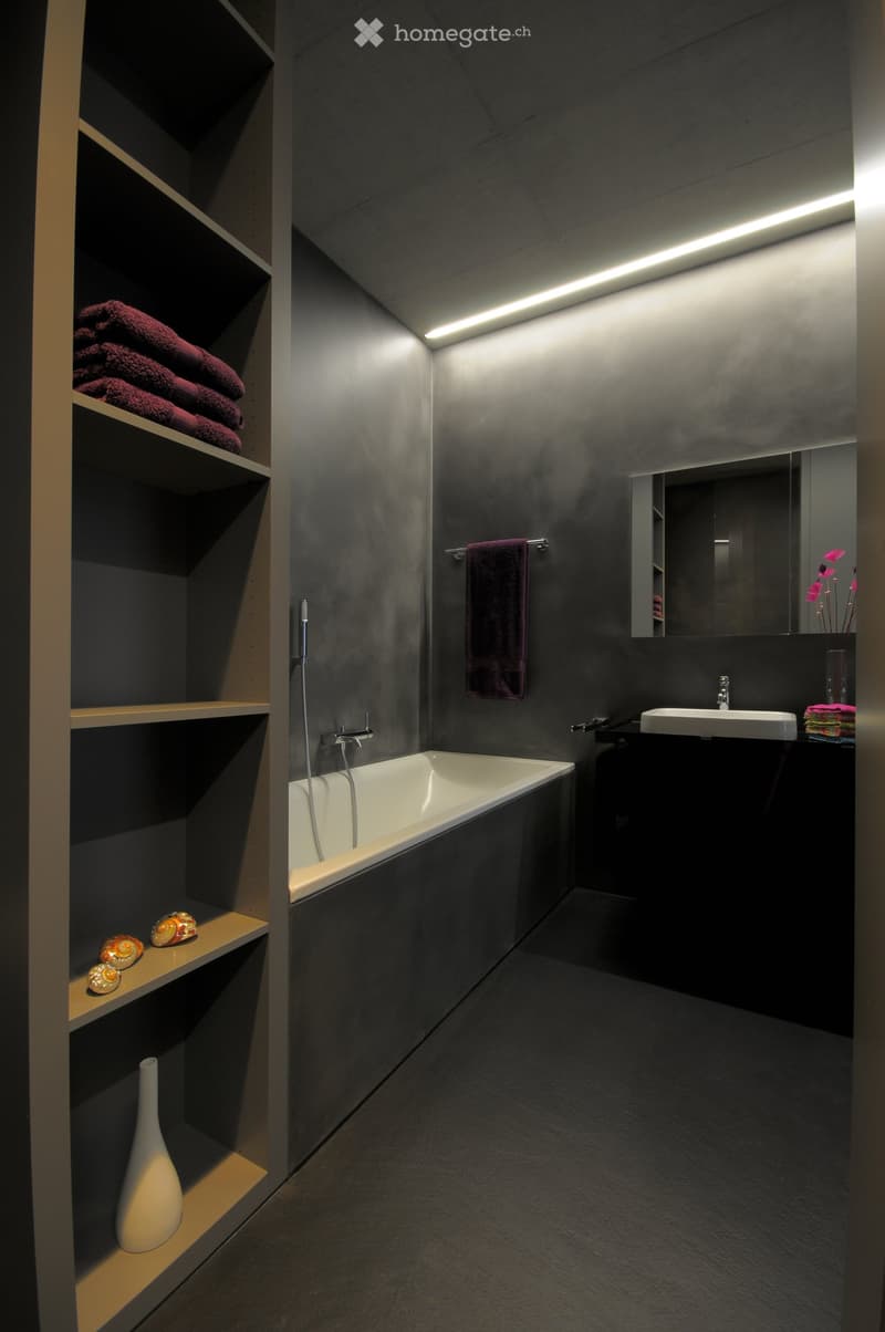Modern bathroom with dark grey walls, a white bathtub, a sink with a mirror, and built-in shelving holding towels and decorative items.