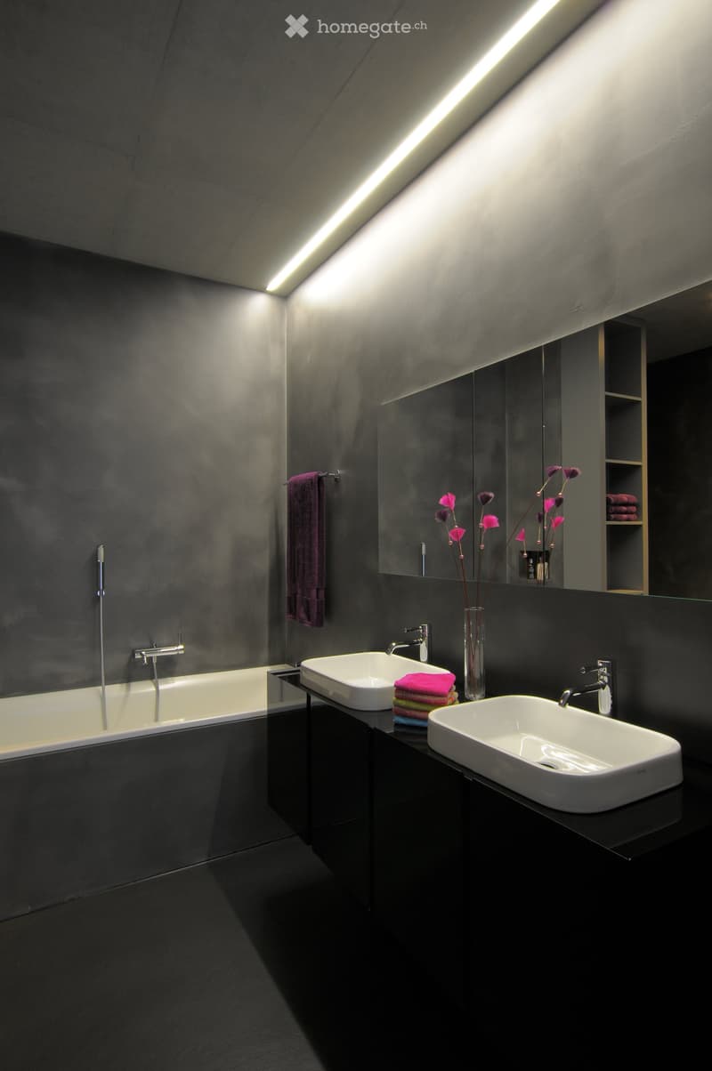 Modern bathroom with dark grey walls and black floor, featuring a bathtub, two white sinks, a large mirror, and a vase with pink flowers on the counter.