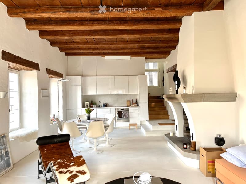 Open-plan living and dining area with a modern kitchen, white dining chairs, a leather sofa, and a fireplace. The space features exposed wooden beams and white walls.