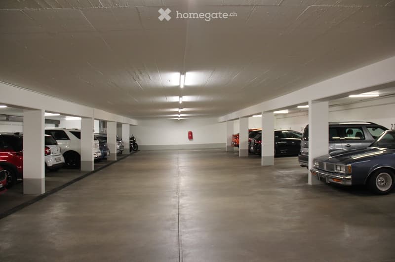 Underground parking garage with multiple parked cars, concrete floors, white pillars, and fluorescent lighting.