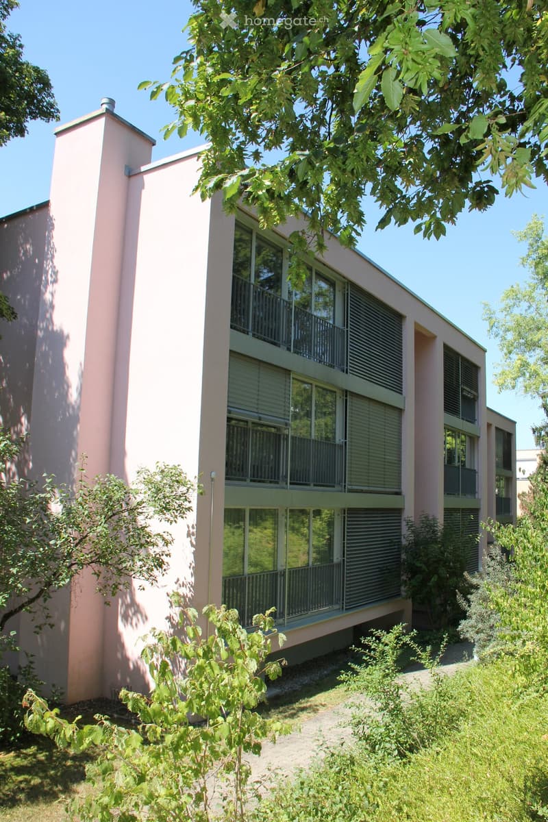 Modern two-story building with pink walls, large windows, and balconies surrounded by greenery and trees.