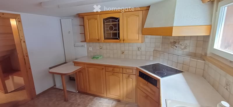 A small kitchen with wooden cabinets, a white countertop, and a built-in electric stove. The space includes a sink, a faucet, and a small table with a green cloth on it.