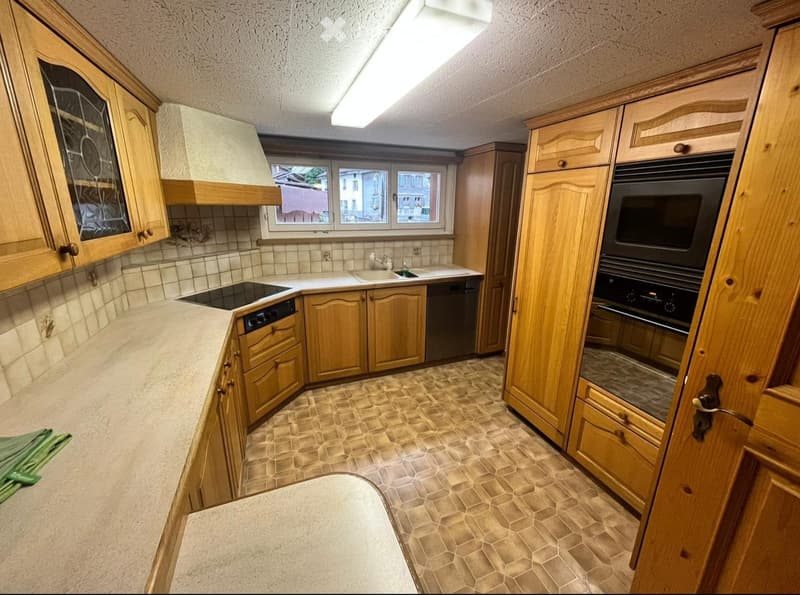 Well-lit kitchen with wooden cabinets, a central island, and modern appliances including an oven, dishwasher, and induction cooktop. The space features tiled walls and a tiled floor.
