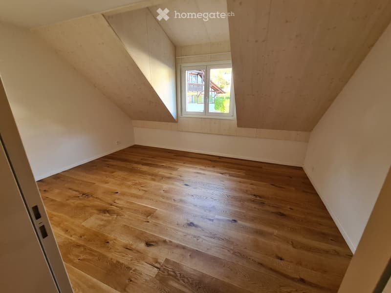 Empty attic room with wooden flooring, white walls, and a skylight window offering a view of the garden and house exterior.