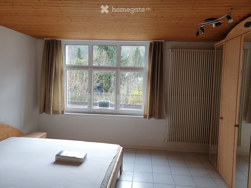 Bright bedroom with wooden ceiling, tiled floor, large windows with curtains, bed, radiator, and wardrobe.
