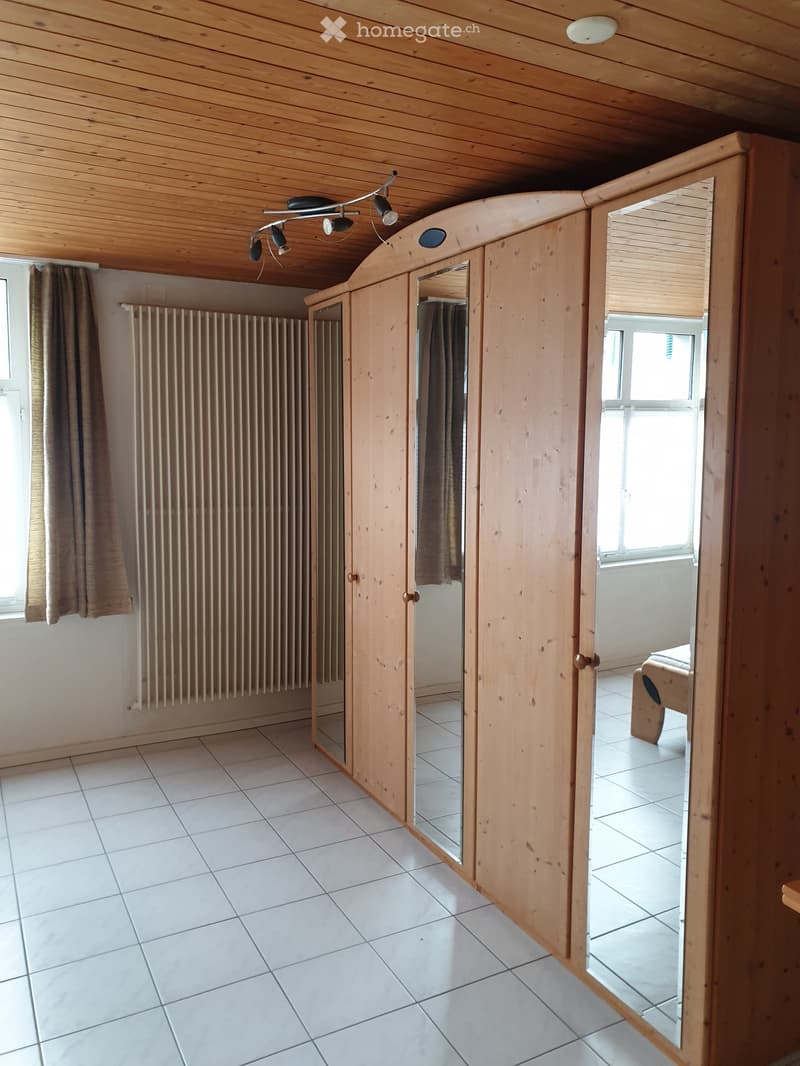 A well-lit room with white tiled flooring, wooden ceiling, and a large mirrored wardrobe. A radiator is visible on the left wall, and a small bed is partially visible in the adjacent room.