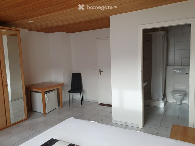 A furnished room with a bed, a small table, a chair, a refrigerator, a mirror, and an adjacent bathroom with a toilet.