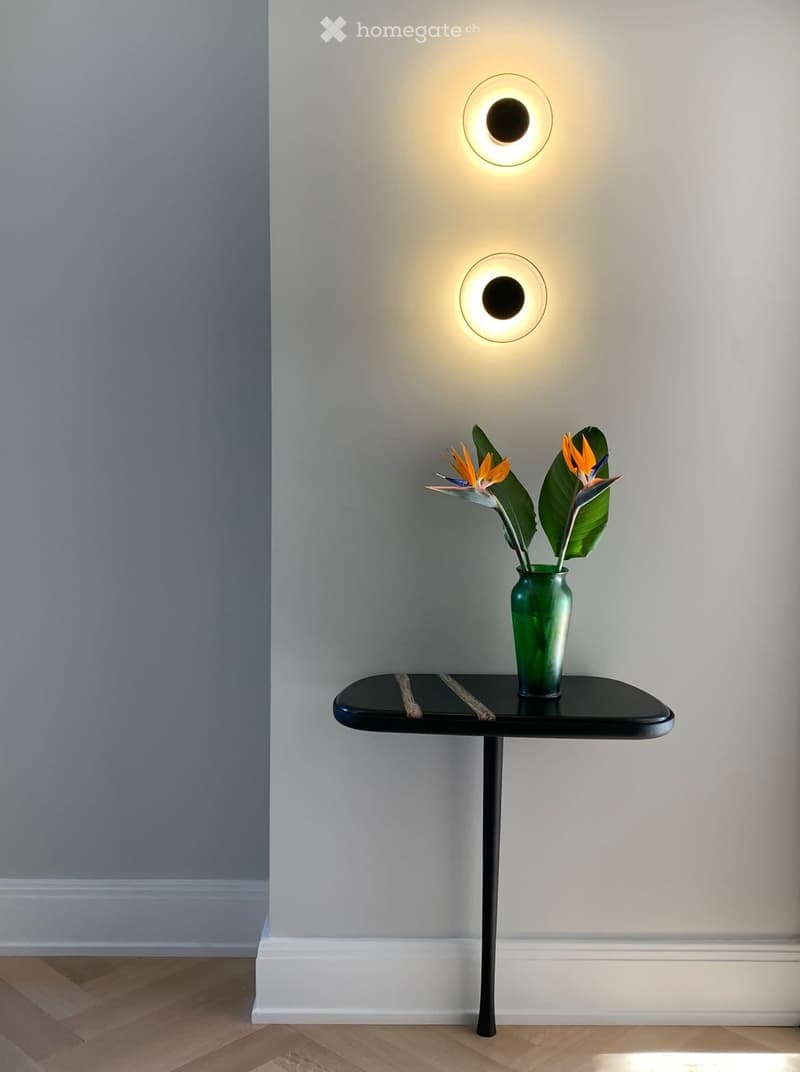 The image shows a minimalist interior design with a black side table or console table, a green vase with colorful tropical flowers, and two circular wall lights providing warm lighting. The background is a neutral gray wall, creating a clean and modern ae