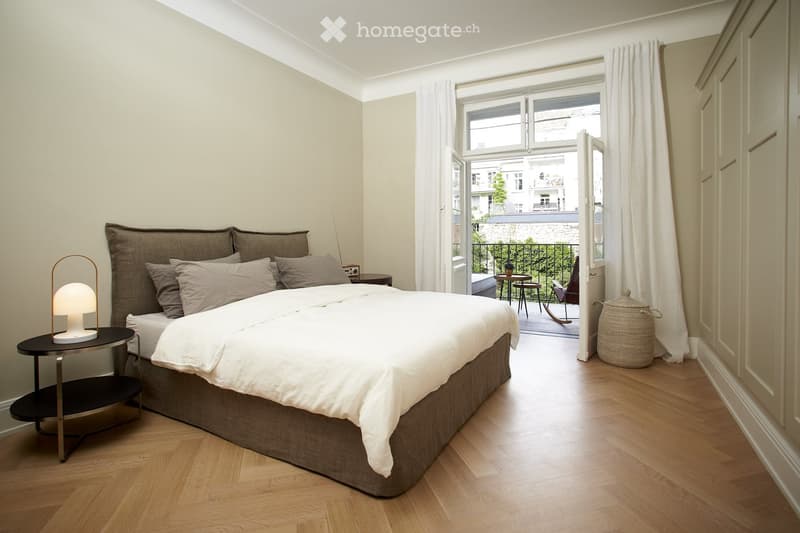 Spacious bedroom with large windows overlooking a balcony, hardwood floors, a king-size bed with a brown upholstered frame, and a side table with a lamp.