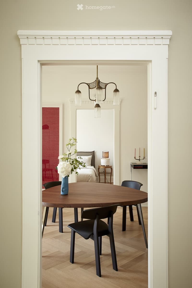 Dining room with a round wooden table, black chairs, a chandelier, and a fireplace visible in the background.