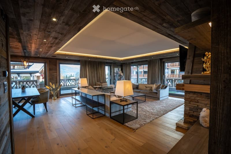 Modern living room with wooden beams, large windows offering a mountain view, a stone fireplace, a sofa, coffee table, armchairs, and a dining area with a table and chairs.