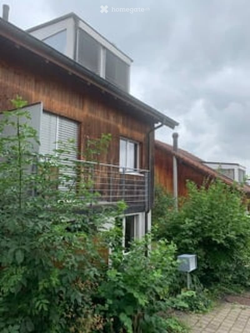 A two-story wooden house with a modern design, featuring large windows, a balcony, and a metal railing. The building is surrounded by greenery and has a paved pathway leading to the entrance.