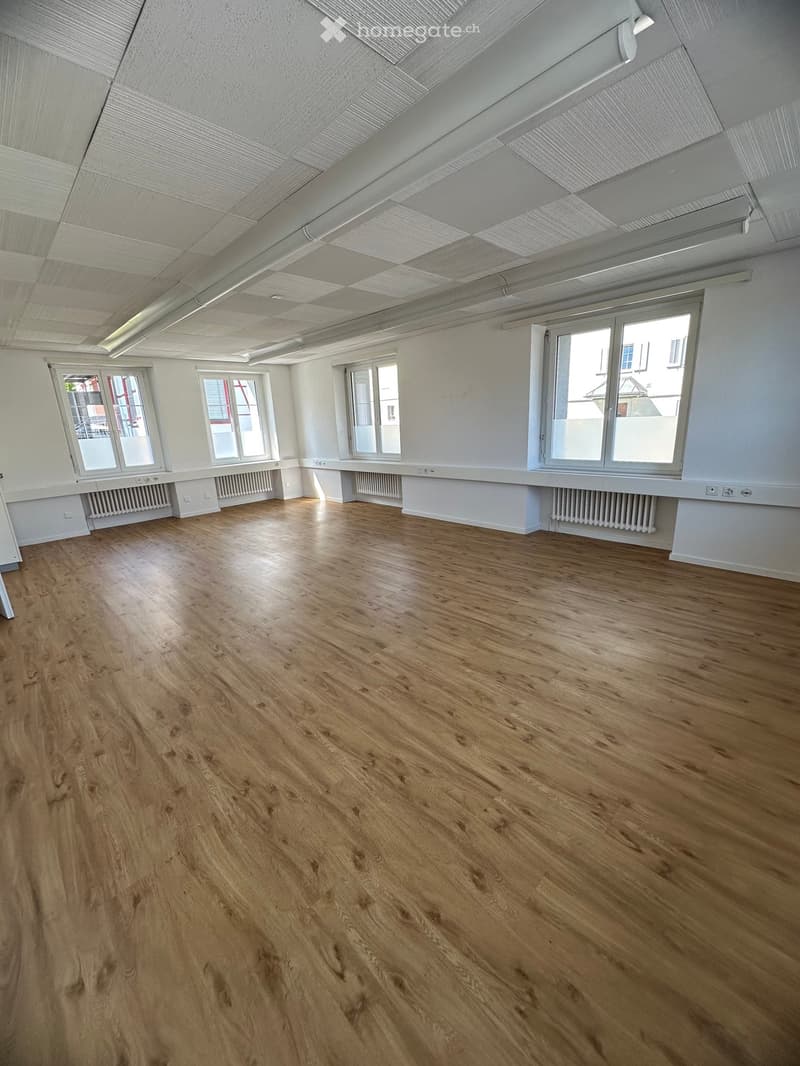 Empty room with wooden flooring, white walls, and large windows allowing natural light. No furniture or equipment is present.