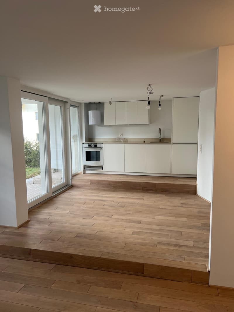 Empty kitchen with light wooden flooring, white cabinets, and a built-in oven. Large glass sliding doors lead to an outdoor area.