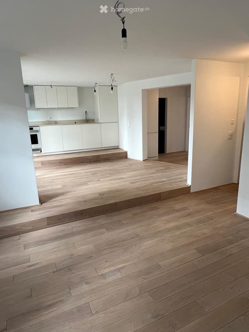 Empty room with wooden flooring, white walls, and visible kitchen area with cabinets and appliances in the background.