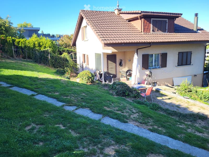 A two-story house with a tiled roof, surrounded by a well-maintained garden with a stone pathway, lawn, and outdoor seating area with a table and chairs.