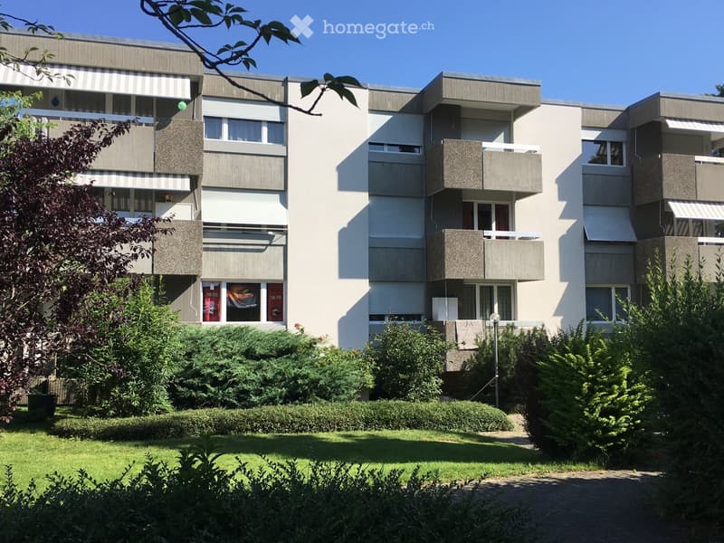 Modern multi-story apartment building with balconies, surrounded by greenery and a well-maintained lawn.