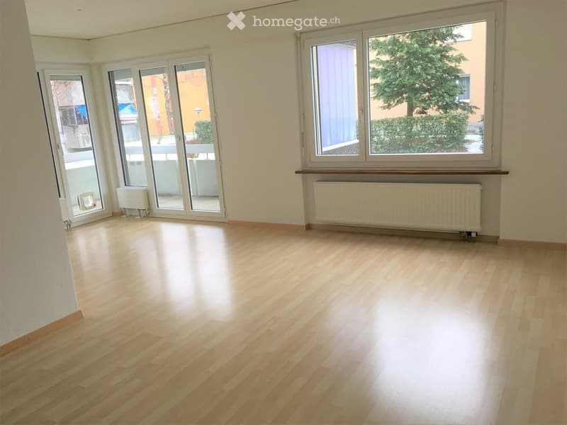 Empty room with light wooden flooring, large windows, and sliding glass doors leading to a balcony area.