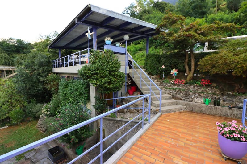 A terraced garden with brick patio, potted plants, stairs leading up to a covered balcony area, and surrounding greenery.
