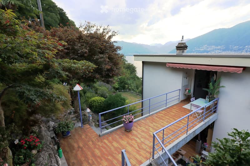 Balcony with brick flooring, metal railing, and outdoor furniture including a table and chairs. Potted plants and a scenic mountain view are visible.