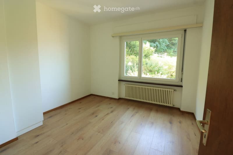 Empty room with light wooden flooring, white walls, and a large window with a radiator below it. The room has a simple, uncluttered design with no furniture or decorations.