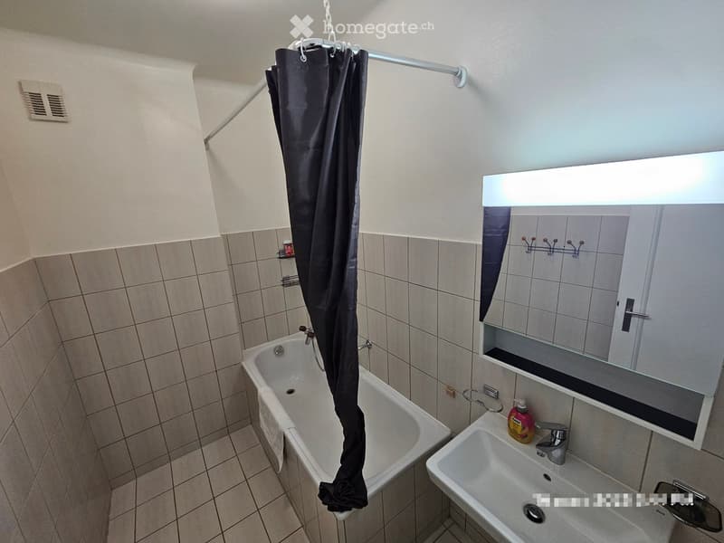 Bathroom with a bathtub, sink, mirror, and shower curtain. The walls are tiled, and a bottle is placed on the sink counter.