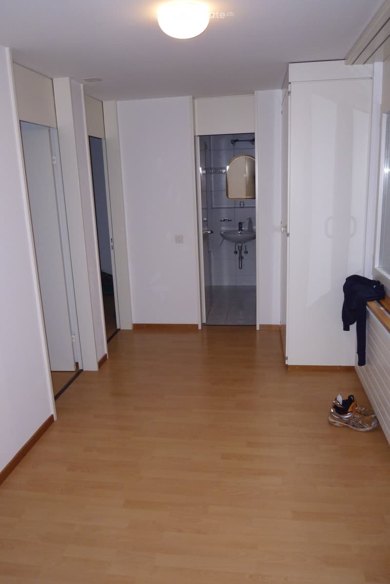 A clean, well-lit hallway with wooden flooring and white walls. A small bathroom is visible at the end, and a pair of shoes and a jacket are placed near the wall.