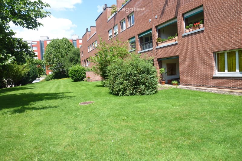 Well-maintained lawn with trimmed grass, surrounded by trees and shrubs, with a brick building in the background featuring balconies and potted plants.