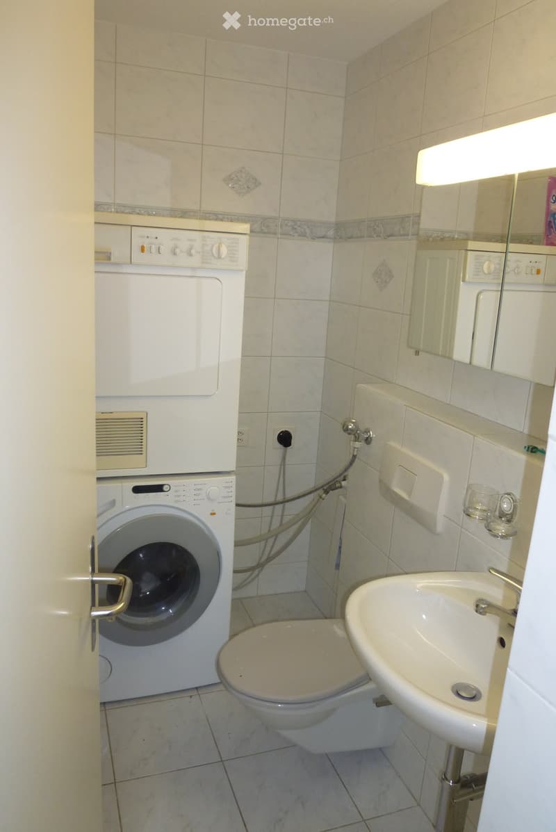 Bathroom with white tiled walls and floor, featuring a toilet, sink, washing machine, and mirror.