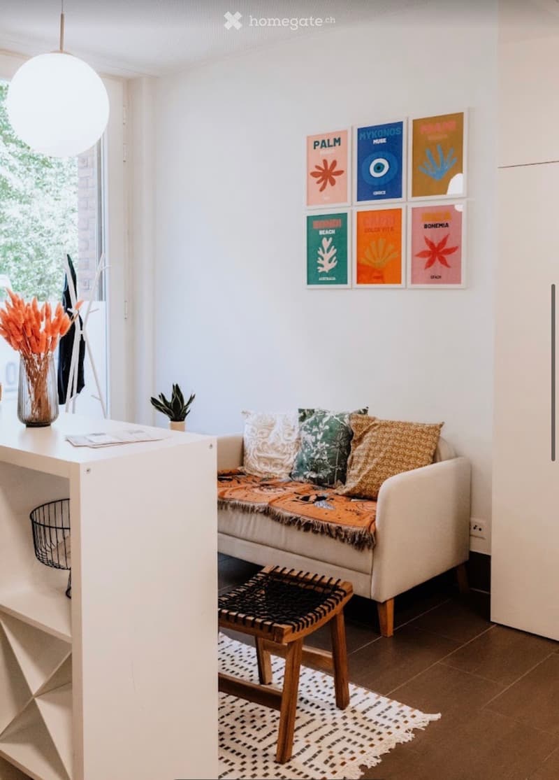 Modern living room with a white sofa, decorative pillows, a small wooden stool, a white table with a vase of orange flowers, and several picture frames on the wall.