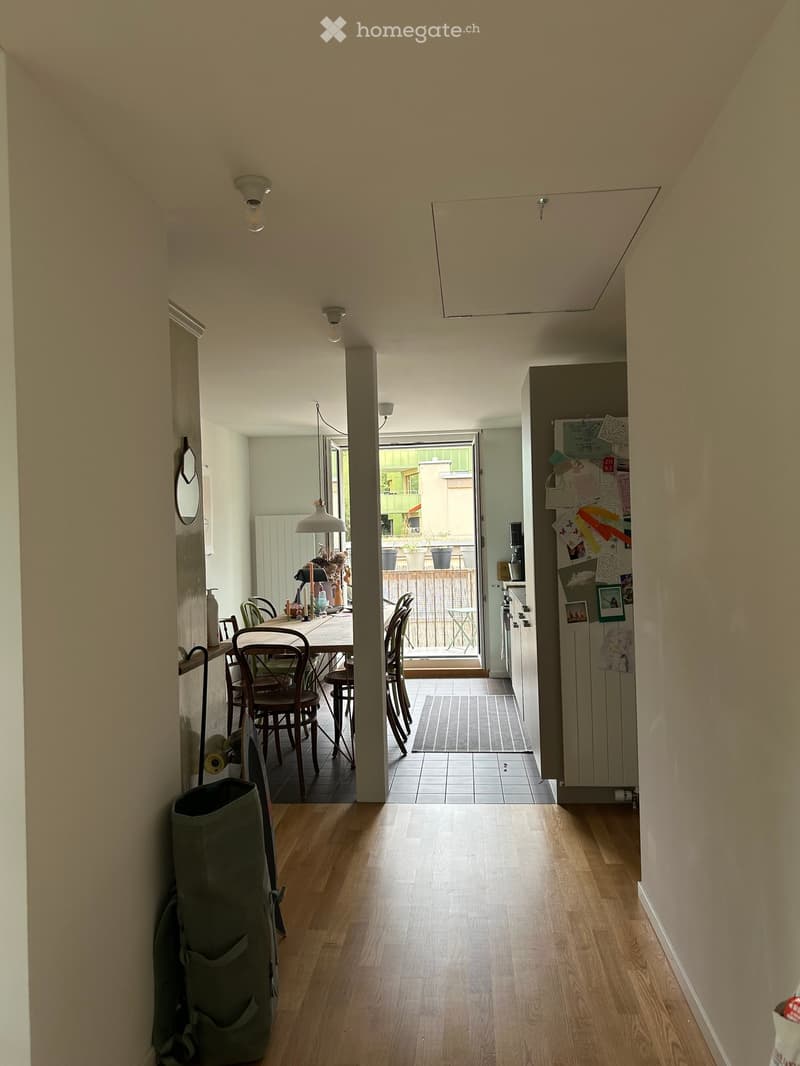 A well-lit hallway with wooden flooring, leading to a dining area and kitchen. The hallway features a trash bin on the left and a mirror on the wall. The dining area includes a table and chairs, and the kitchen has a refrigerator with notes attached.