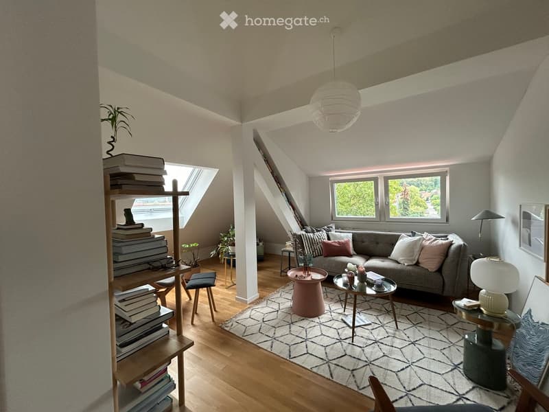Cozy living room with wooden floors, large windows, a grey sofa with pillows, a coffee table, side tables, a bookshelf filled with books, and a patterned rug.