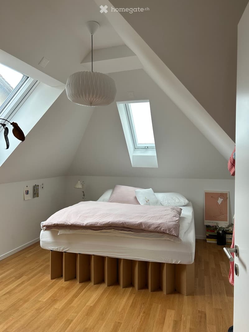 Cozy attic bedroom with wooden floors, slanted ceilings, and skylights. Features a bed with pink bedding, a pendant light, wall lamp, picture frame, and books on a shelf.